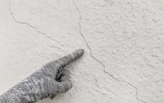 Hand of industrial worker in construction glove points to cracks on white plaster wall texture background
